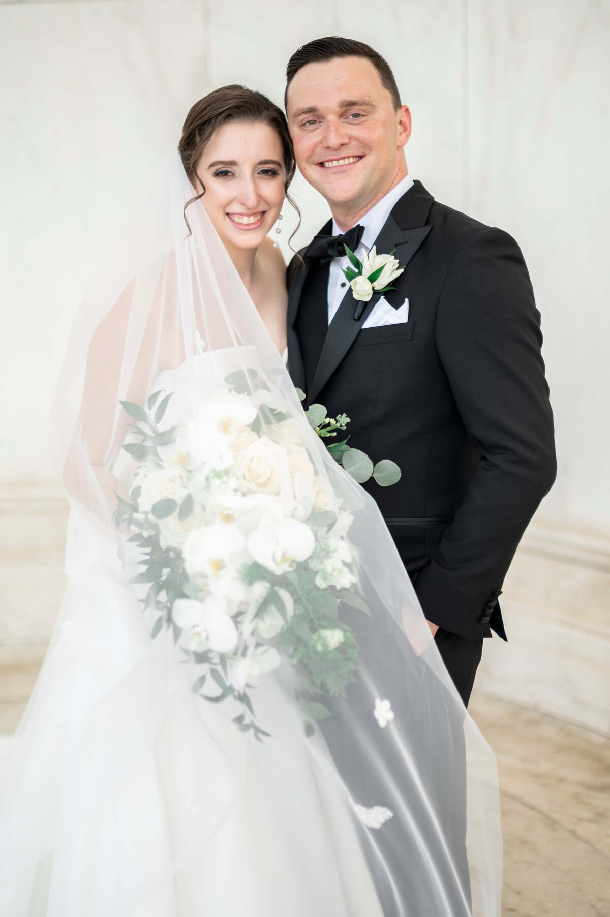 A bride and groom smile happily on their wedding day. The bride wears a white dress with a veil and holds a bouquet of white flowers. The groom is in a black tuxedo with a white boutonnière. They stand close together in a light, elegant setting.