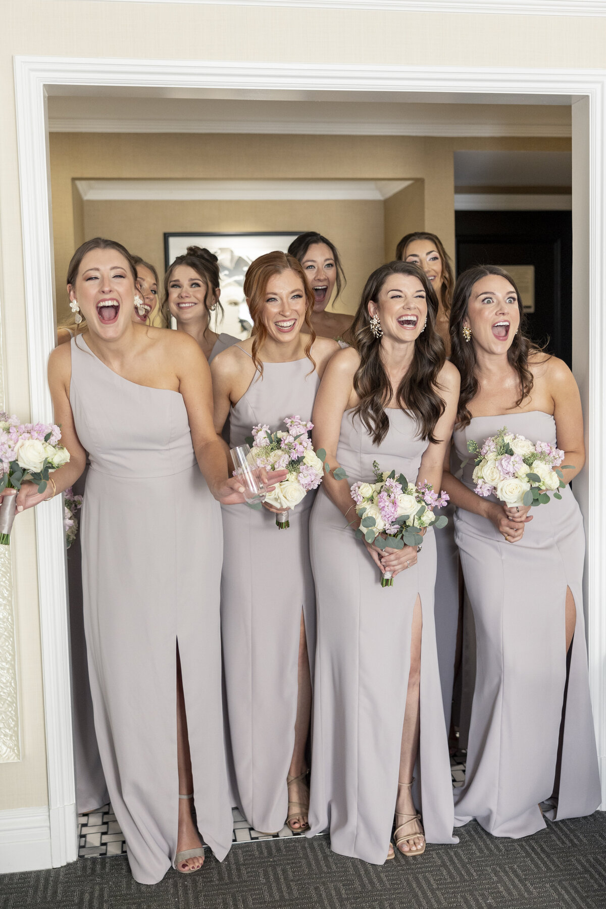 A group of bridesmaids in light lavender dresses stand in a room, holding bouquets of pink and white flowers. They are smiling widely and appear excited.