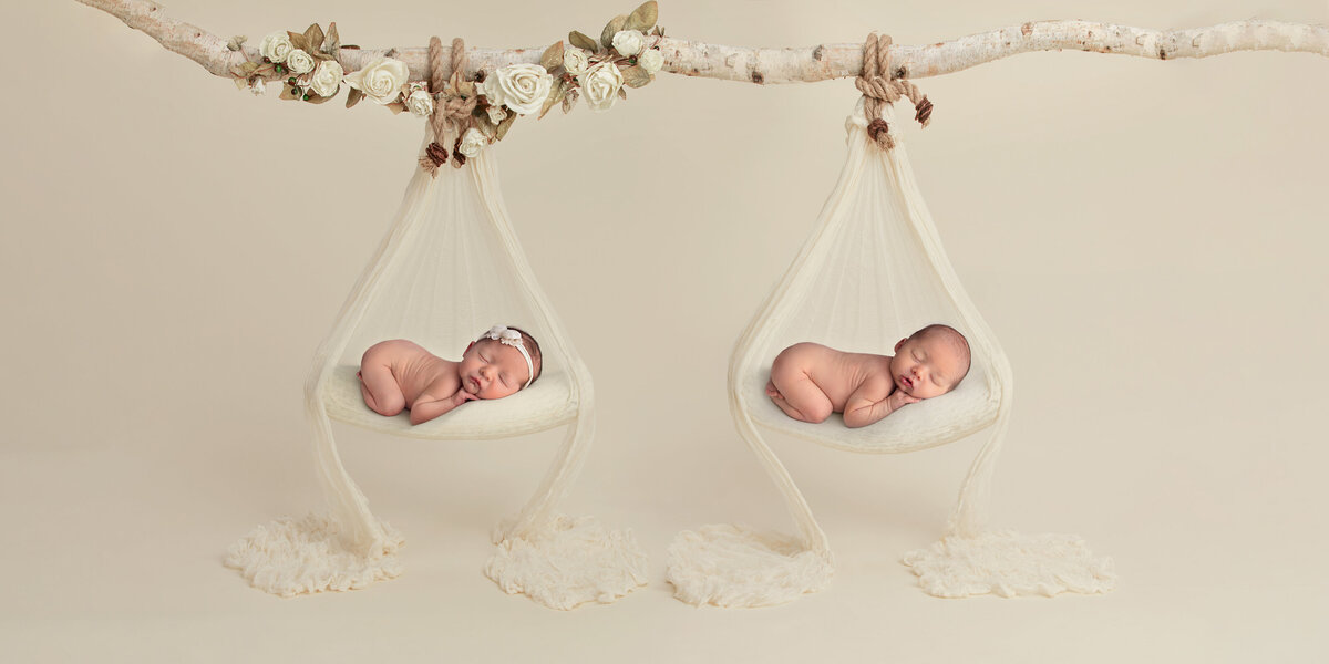 Newborn twins posed individually on cream hanging swings decorated with white flowers, both sleeping peacefully on a neutral background.