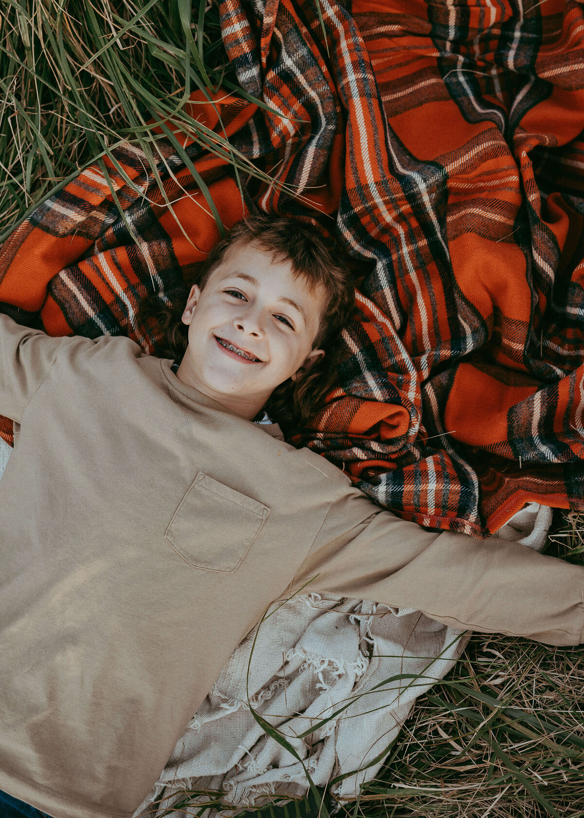 A cute boy with a crazy mullet, wearing a tan color shirt, lays on a vibrant orange plaid blanket on his families farm during a fall outdoor photo session.