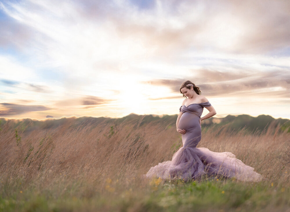 Dramatic sunset maternity photo in Westlake, Texas, showing a mother-to-be in a whimsical lavender gown walking through golden prairie grass.
