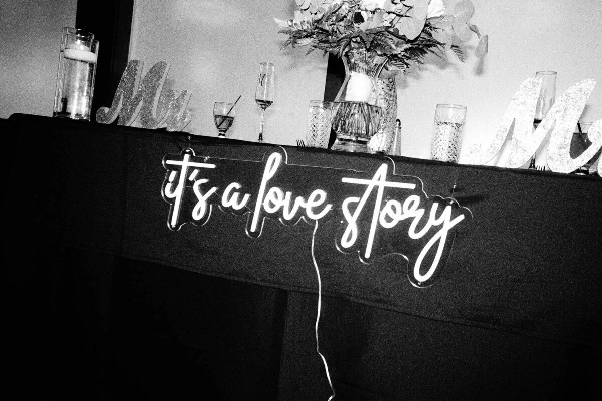 A neon sign reading its a love story hangs on a table, decorated with glasses, a vase of flowers, and Mr and Mrs signs in the background. Captured in black and white by an NJ wedding photographer.