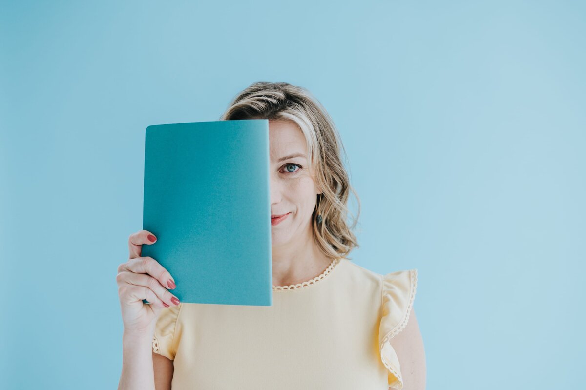 Blonde woman holds aqua book in front of half of her face while smiling to the camera.