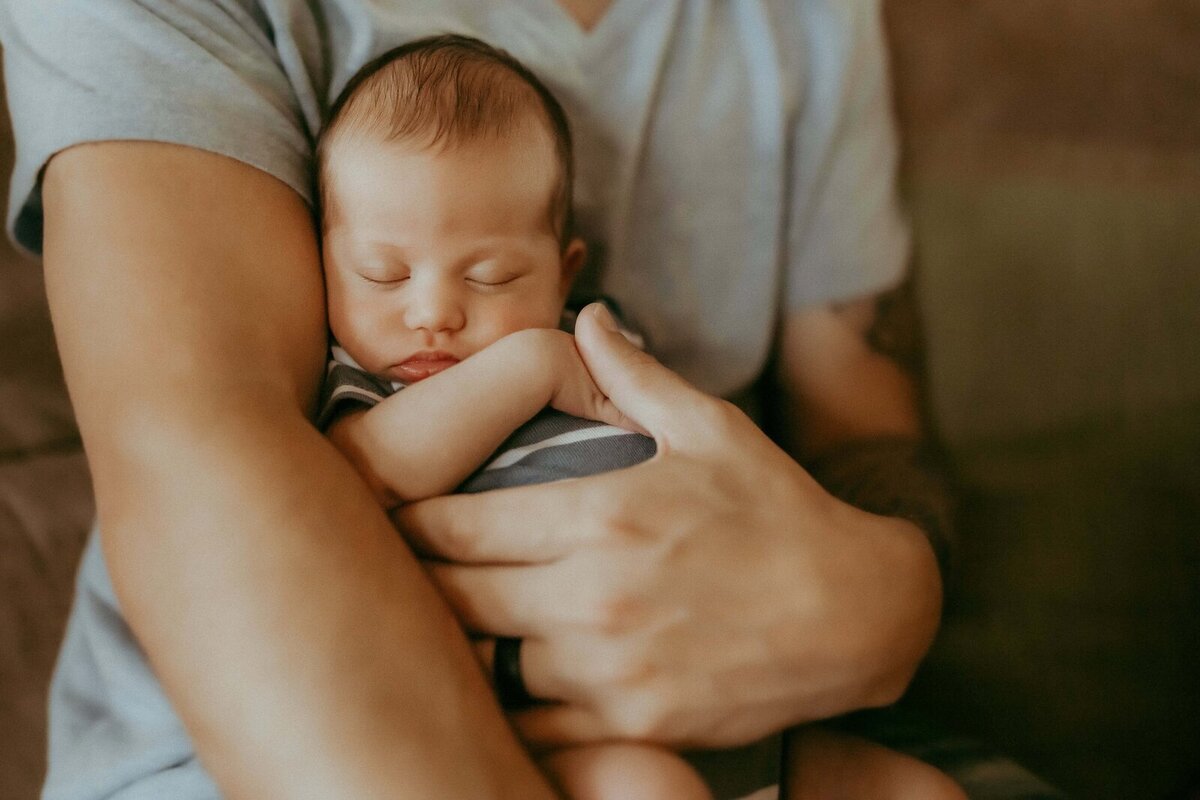 A newborn baby sleeps peacefully in an adult's arms, both dressed in grey clothing, against a blurred background.