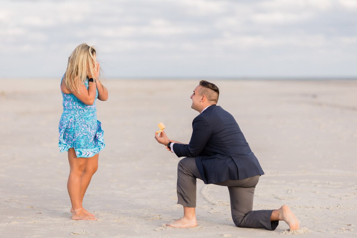 Guy on one knee proposing to a surprised bride to be at the beach by Congress Hall in Cape May