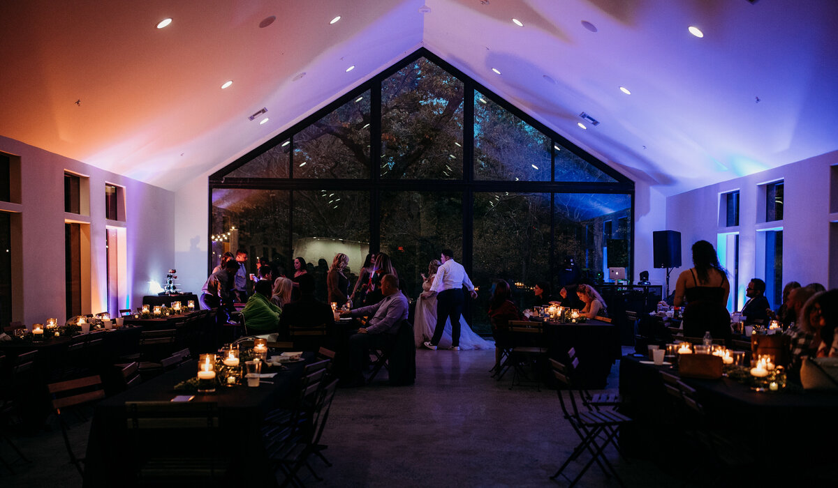 Elegant wedding venue at night with a glass front, dimly lit with purple lighting, and guests mingling at tables
