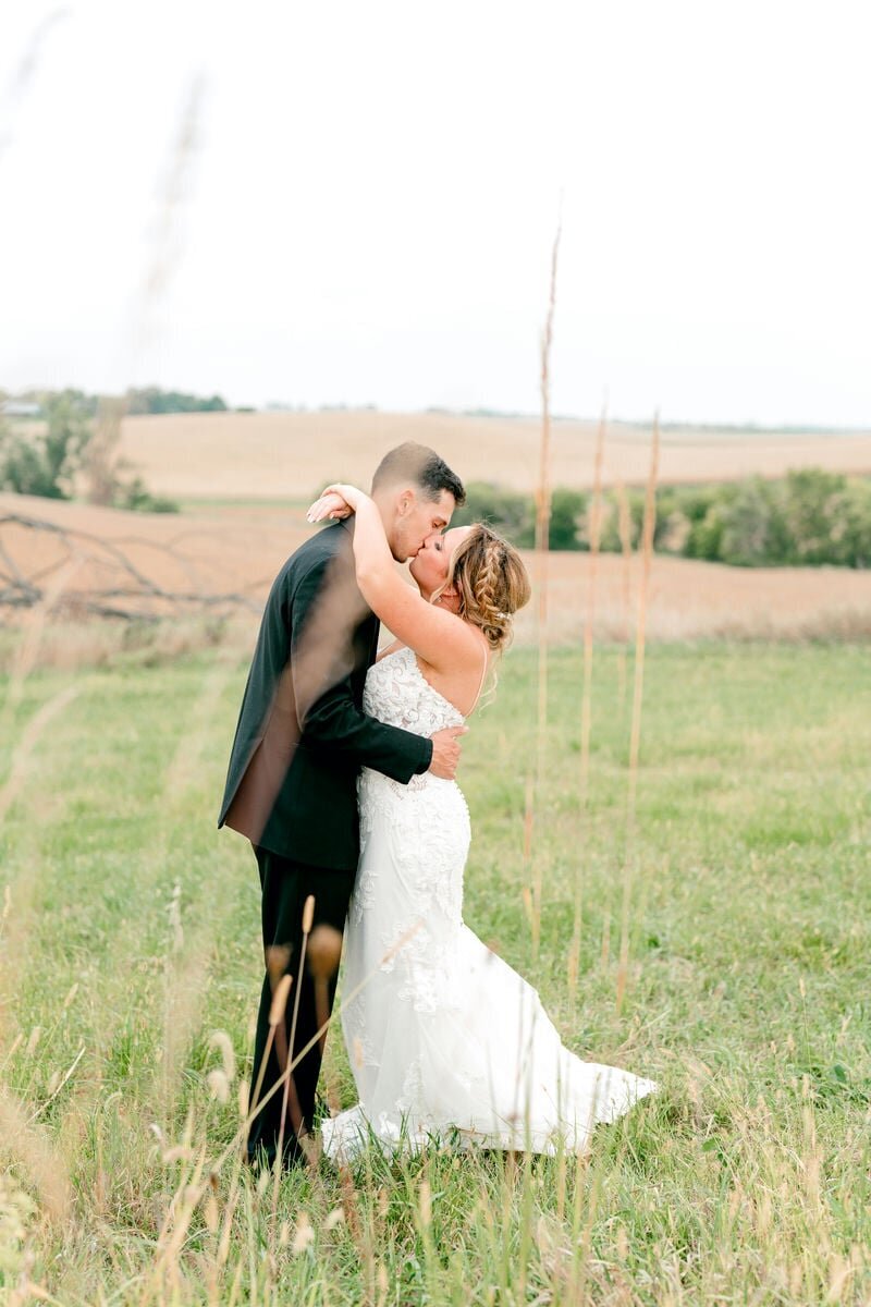 A bride and groom share a romantic kiss in a serene open field, surrounded by tall grasses and a beautiful rural landscape, capturing a tender moment on their wedding day.