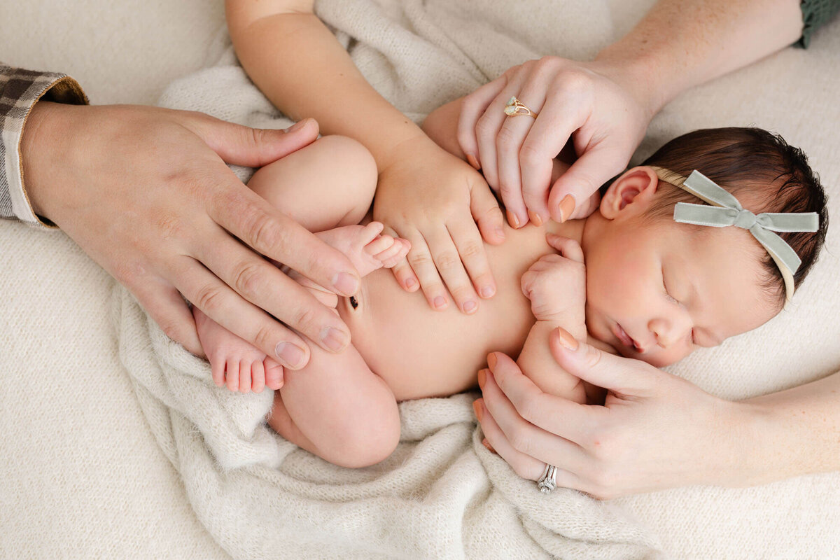Newborn baby laying on textured beige blankets sleeping while mom dad and big brother all rest their hands on baby. Image taken from above, so you are looking down at baby.