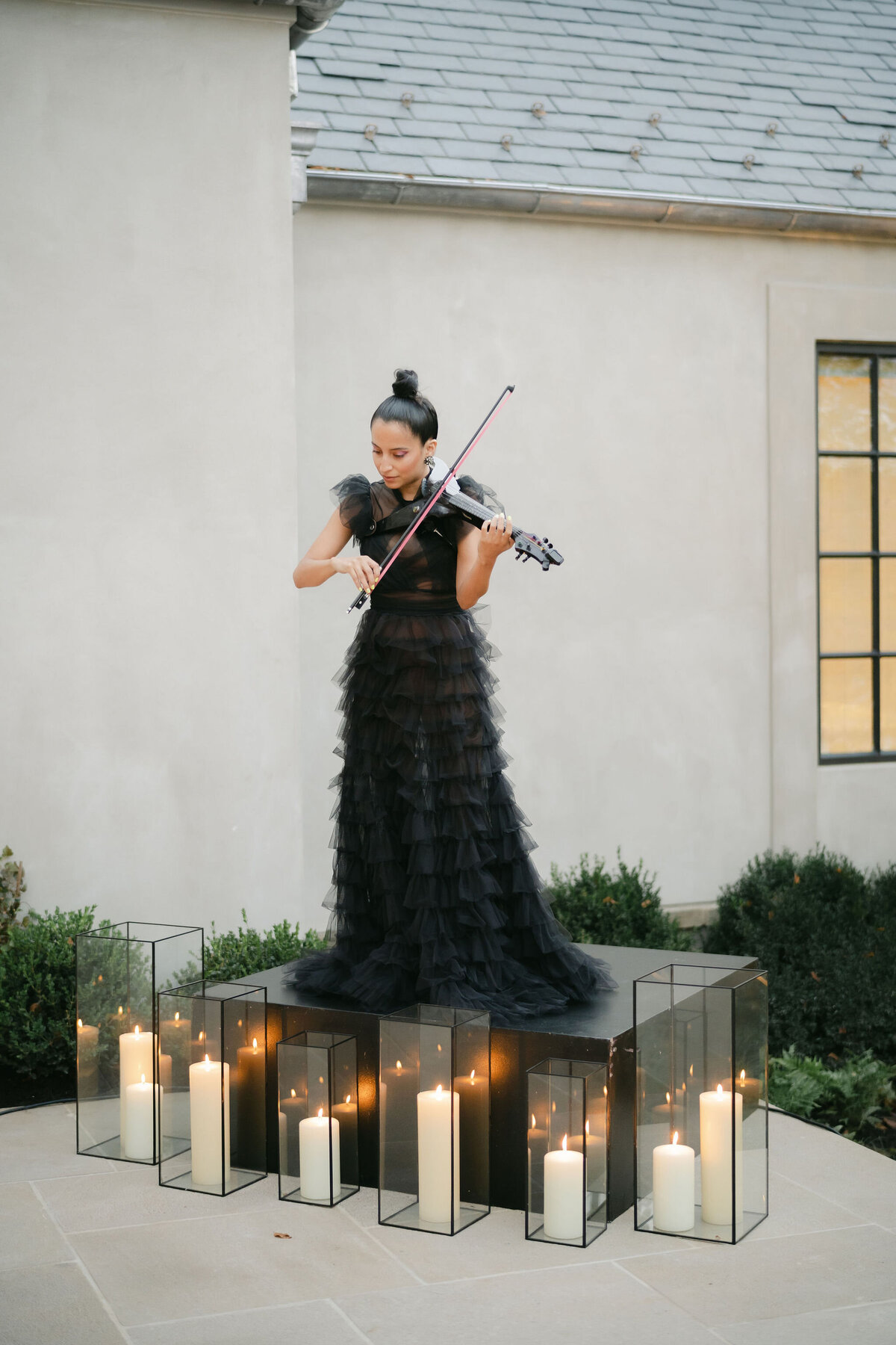 The beautiful photo was taken at a tented wedding in Virginia by the talented photographer Elisabetta Marzetti, with Clayton Films providing videography services. In the picture, a woman dressed in a flowing black gown elegantly plays the violin while standing on a small platform outdoors. Surrounding her are black-framed glass candle holders of varying heights, each holding a flickering candle to create a warm and romantic atmosphere. The minimalist wall and lush plants in the background add a touch of elegance to the scene.