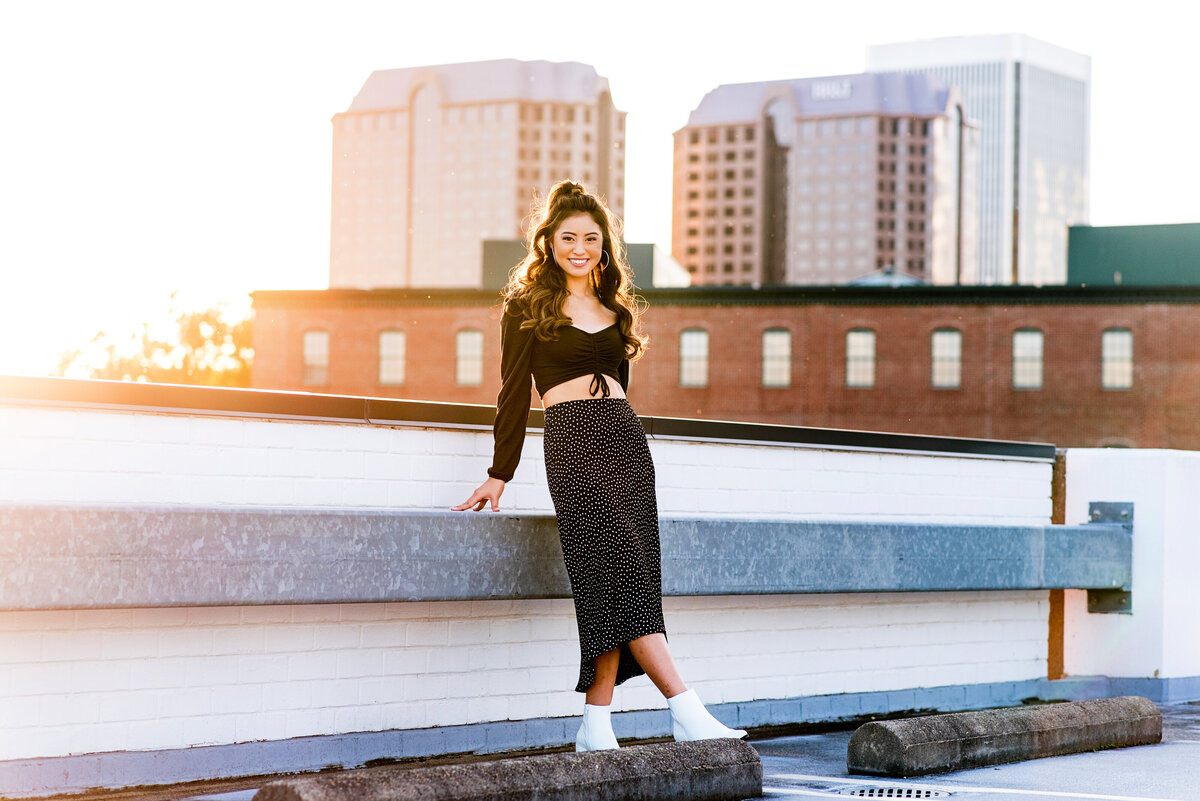 Senior girl poses on rooftop at sunset. View of downtown Richmond in background.