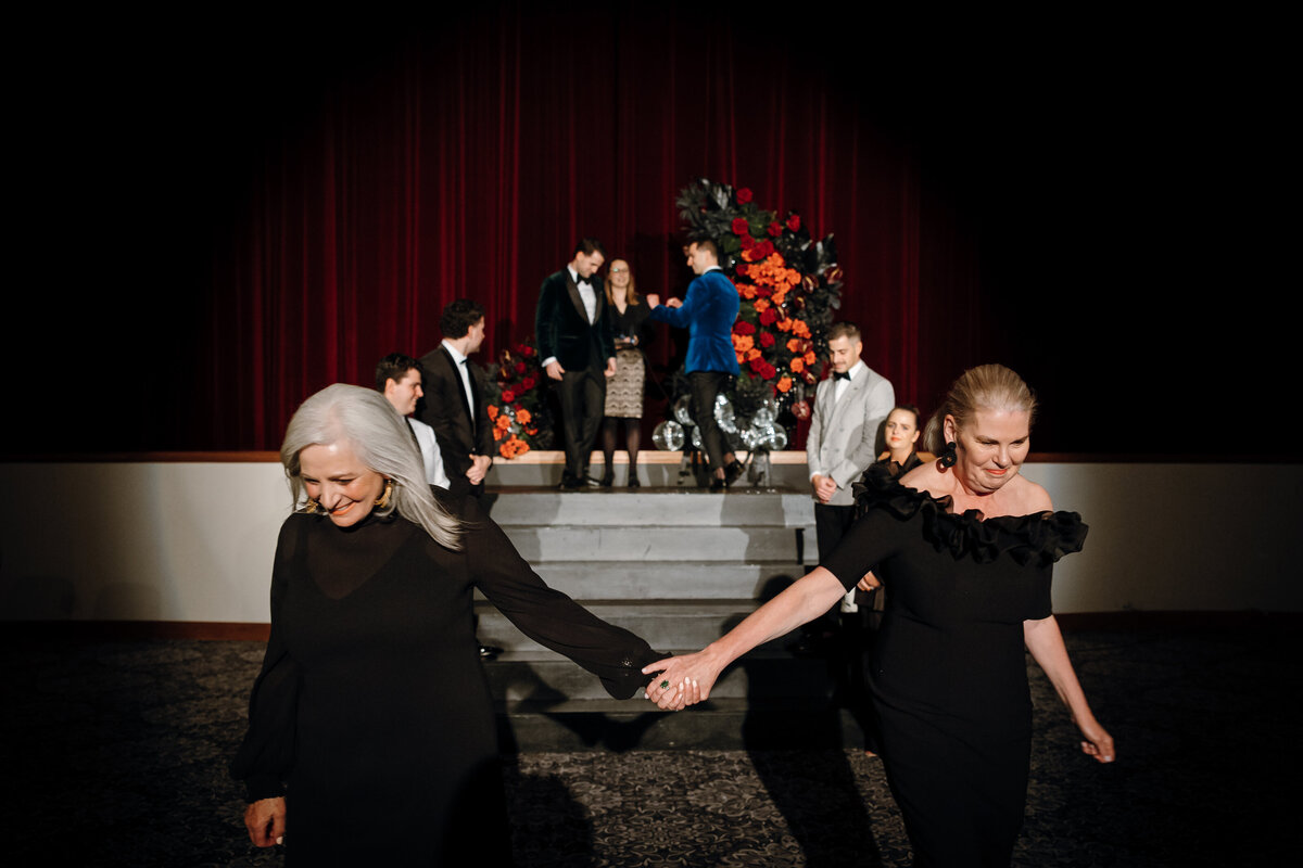 Two women hold hands as the grooms walk up the stage.