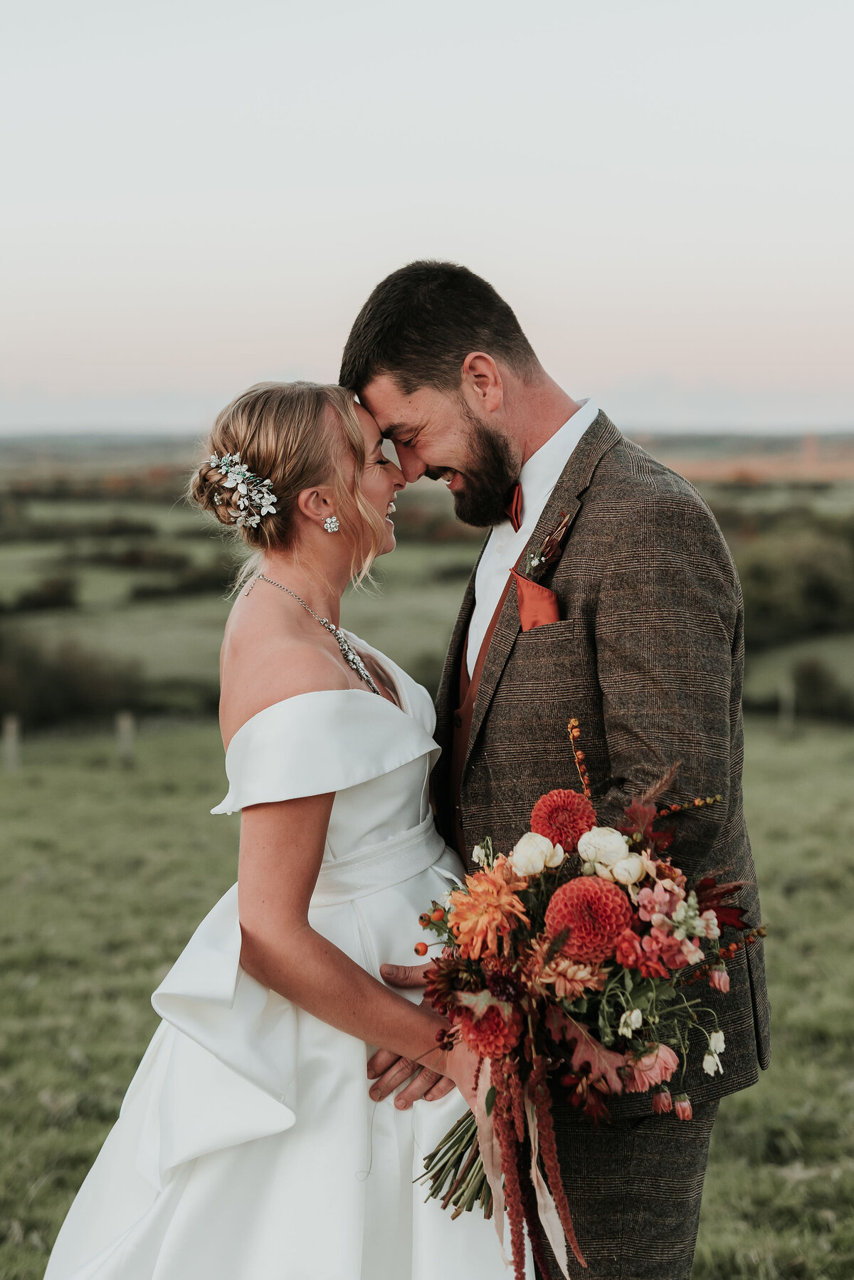 Bride & Groom embrace gently forehand to forehand holding a beautiful Autumn floral bouquet at their rustic fall wedding at Montague Farm, Sussex