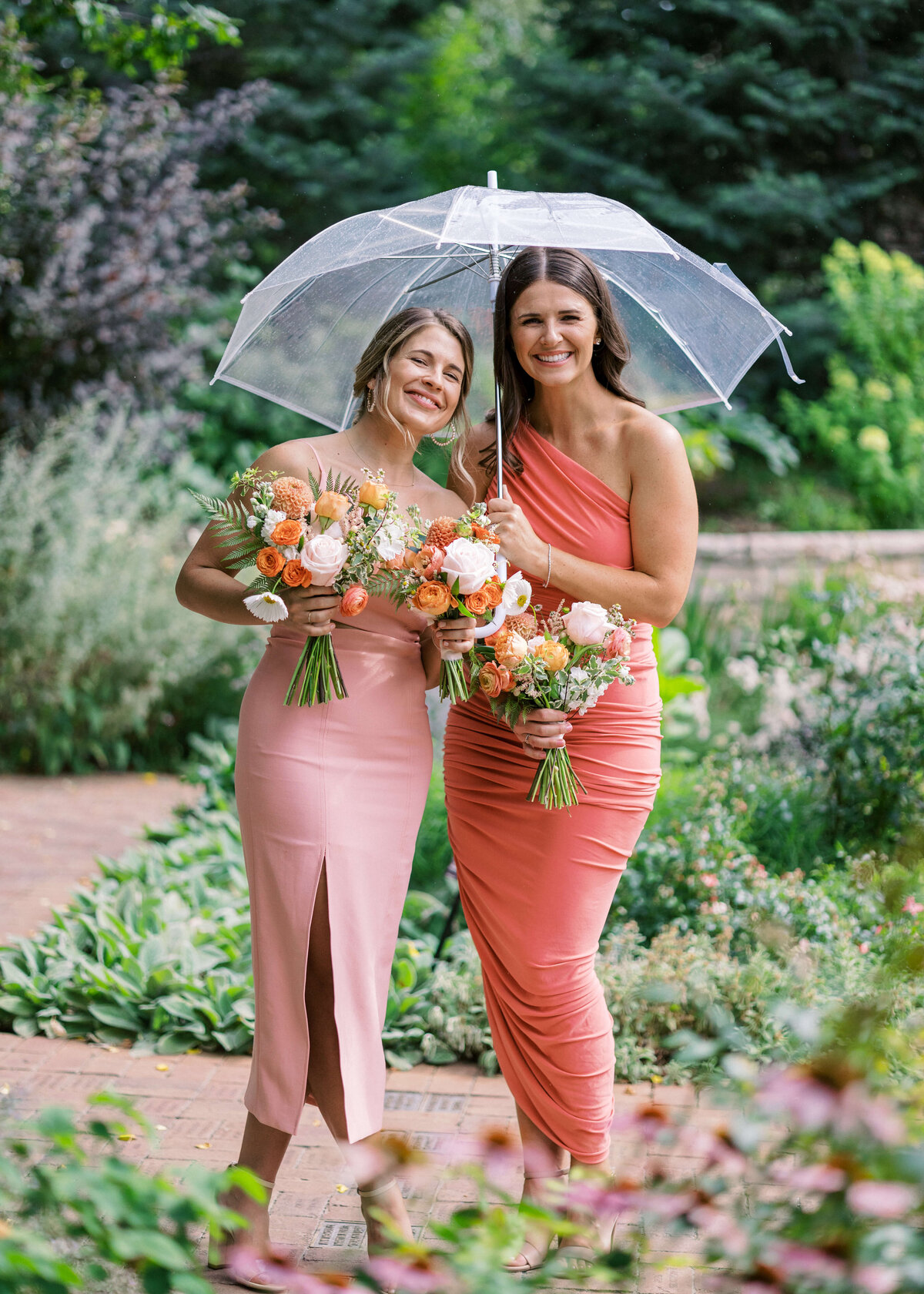 Two bridesmaids in pink and coral dresses hide under a clear umbrella during cocktail hour