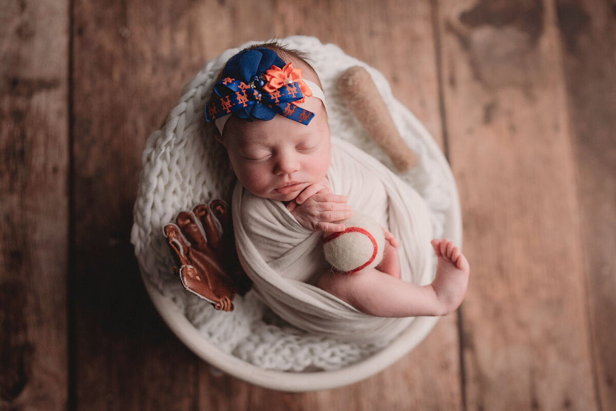 Newborn baby laying in white bowl on wooden floor backdrop baby is holding a baseball