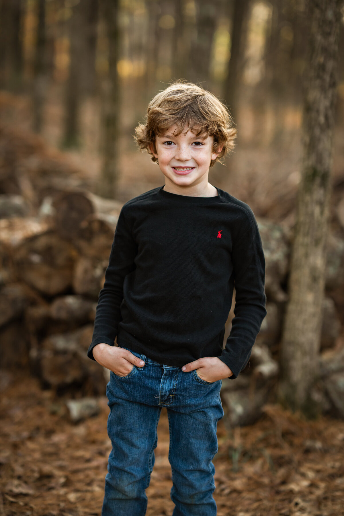 A young boy in a black shirt stands in the woods and smiles for the camera at his photoshoot.