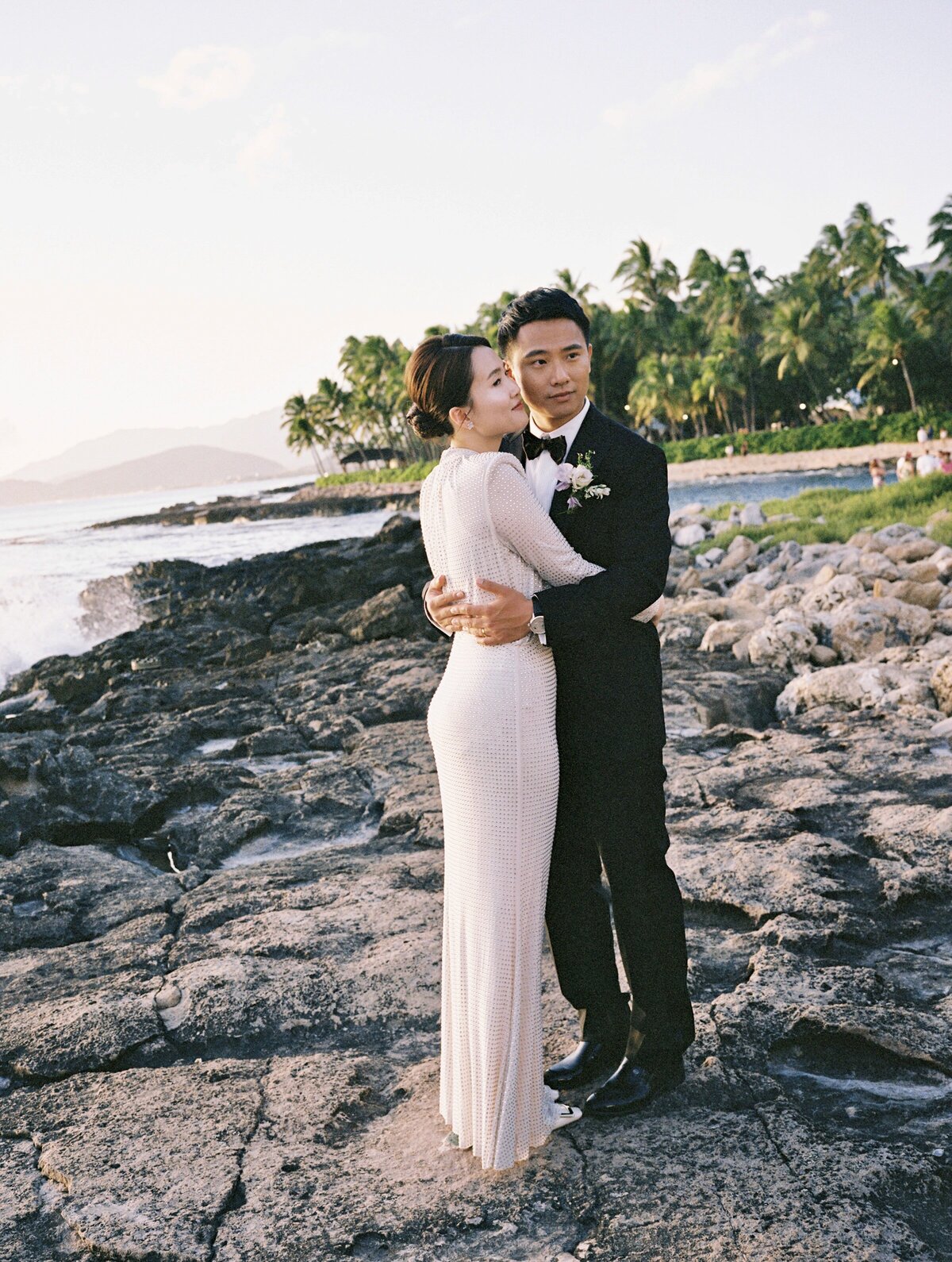 Shy couple embraces each other on the rocks in Oahu, captured in a timeless and romantic moment by Kate Winters.