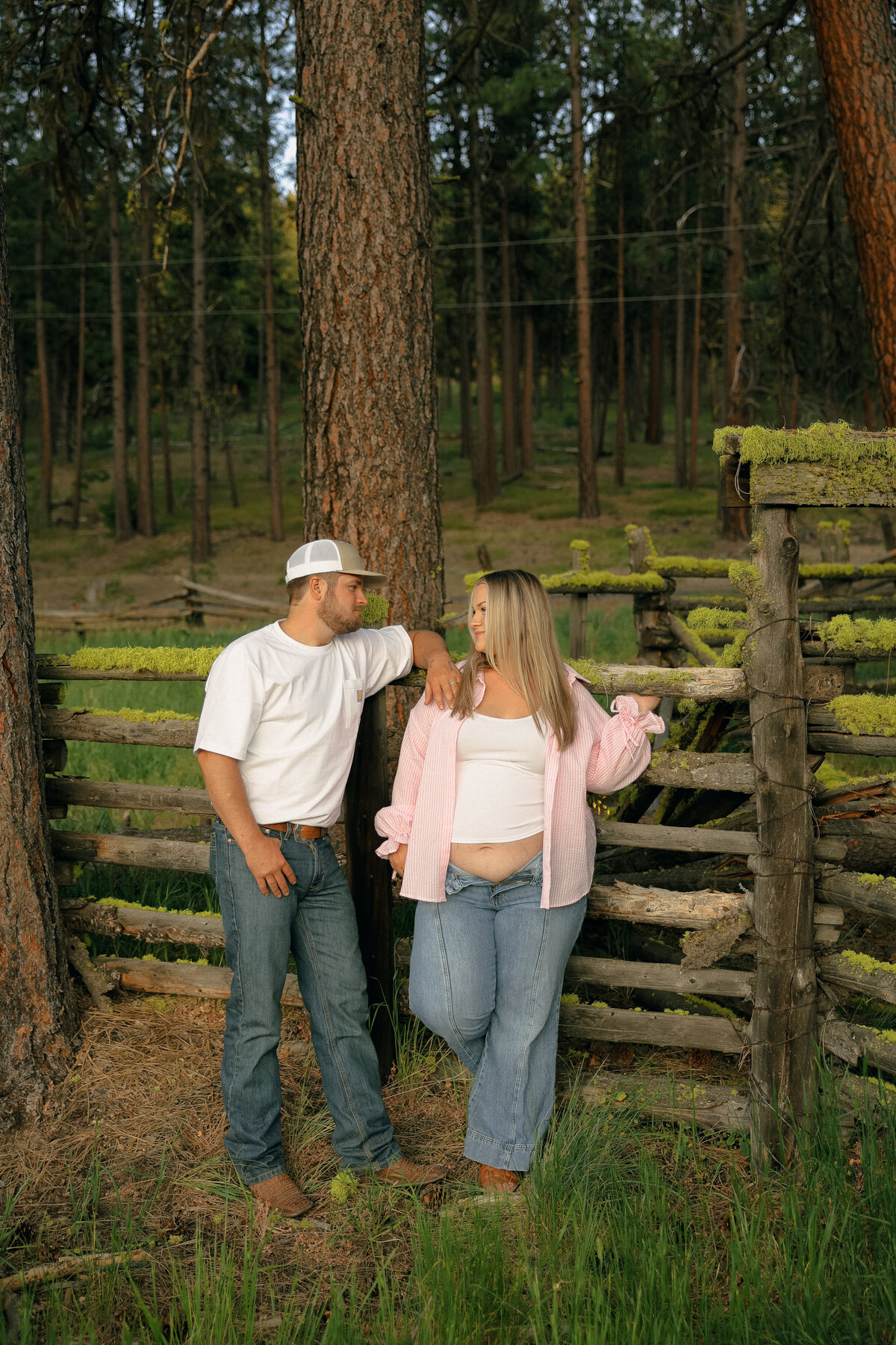 Rustic Maternity Session with Couple Leaning on Wooden Fence Surrounded by Forest Trees