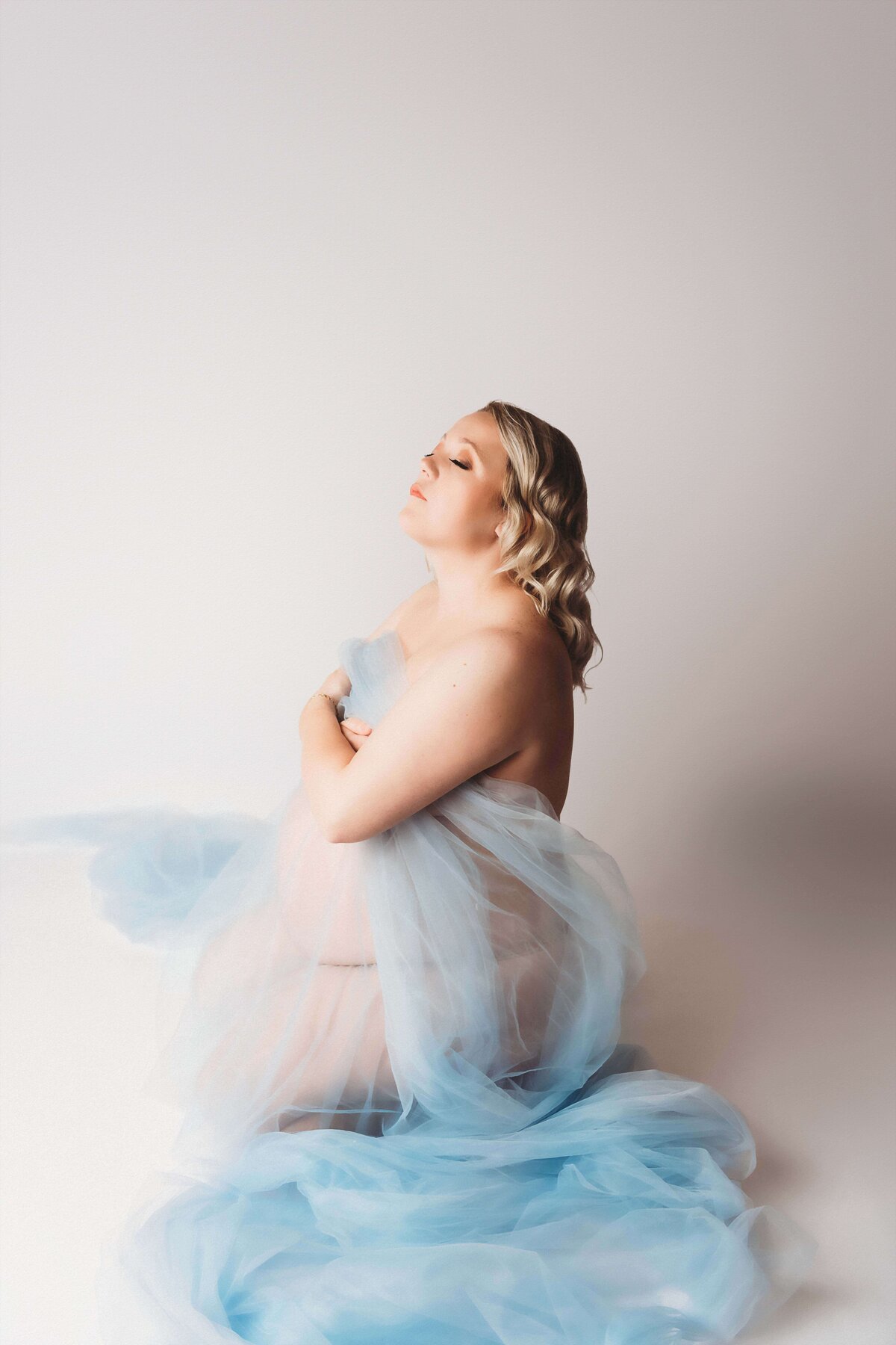 An expecting mama poses on her knees, resting on a white backdrop. She is wrapped in blue tulle to signify the son she is expecting soon.