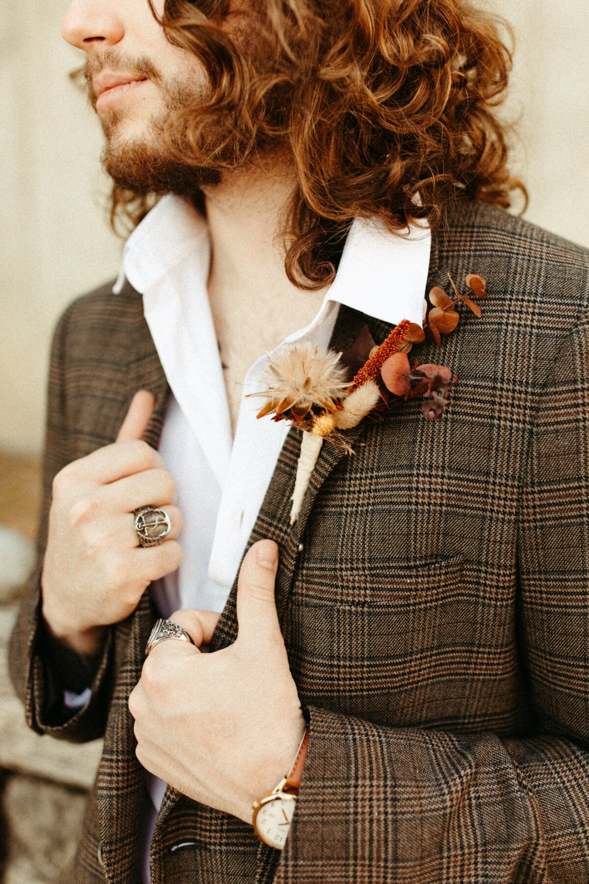 Close up of boho groom's dried floral boutonniere as he adjusts his plaid suit and shows off his rings and waatch