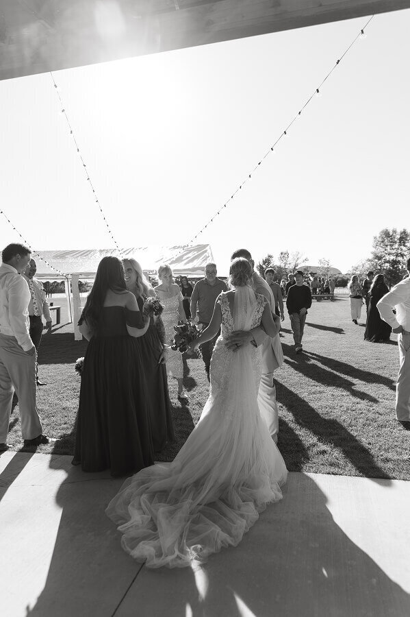 A photo of the groom from behind with his arm around his bride's waist. They're at their outdoor wedding reception mingling with guests.