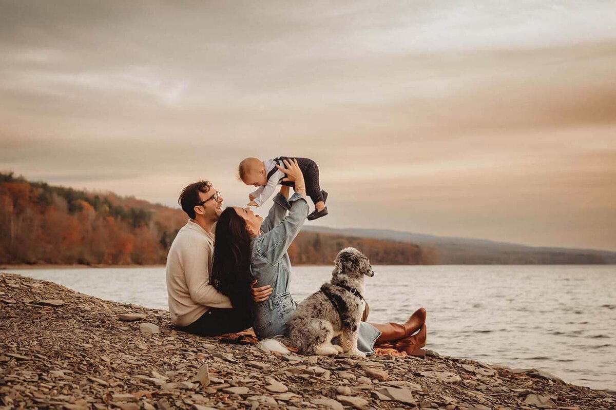 a mom and dad with with their dog on the shore while the mom lifts her baby in the air