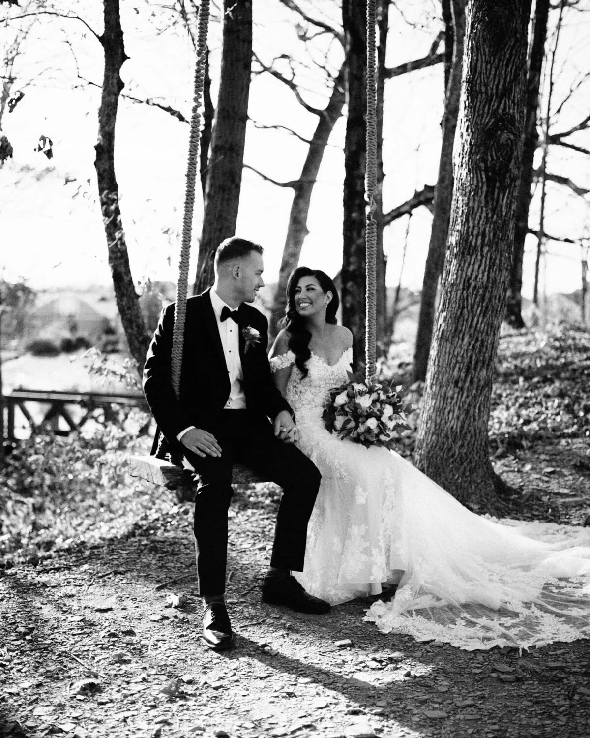 A bride and groom sit together on a swing in a wooded outdoor setting. Captured by an NJ wedding photographer, the bride holds a bouquet and smiles at the groom as sunlight gently filters through the trees.