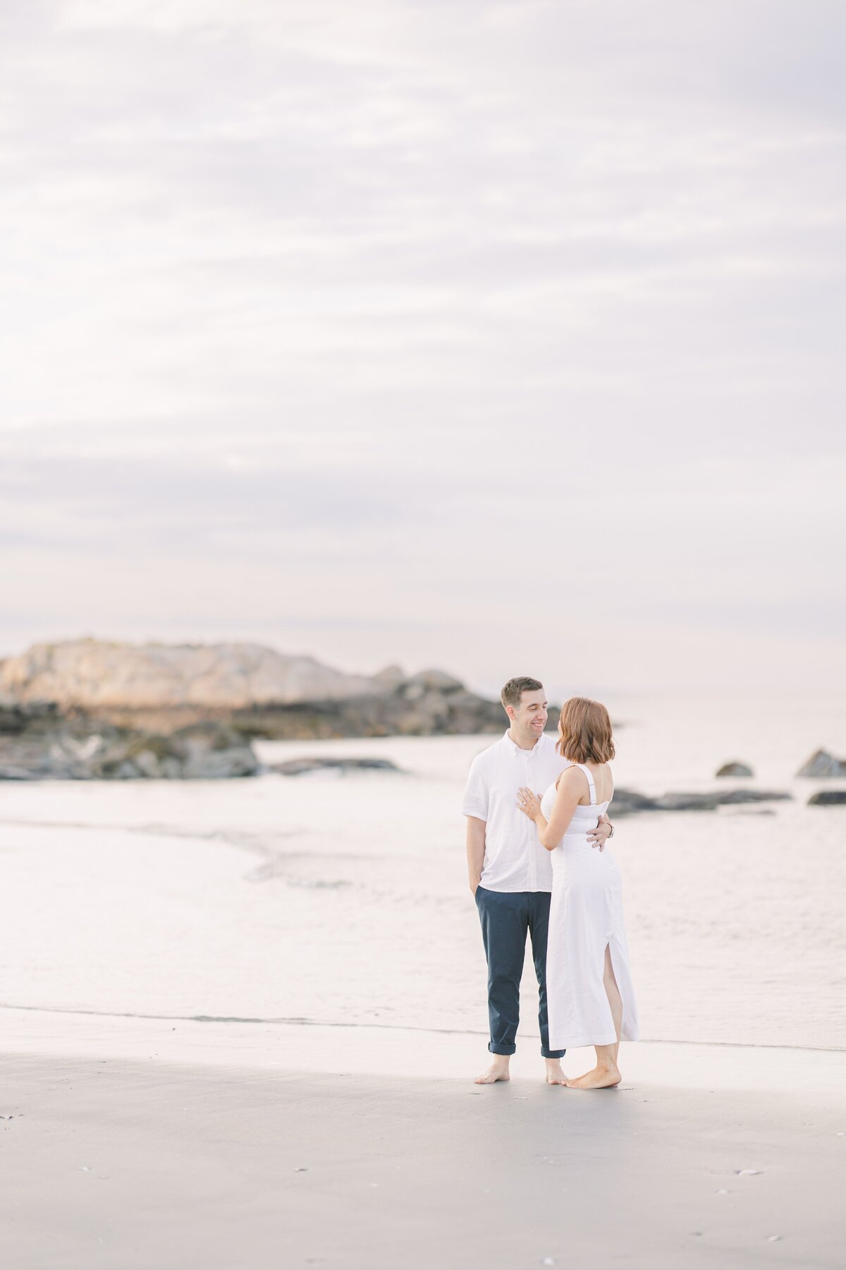 A woman leaning in towards her laughing fiance on Wingaersheek Beach during their engagement session with a Gloucester photographer.