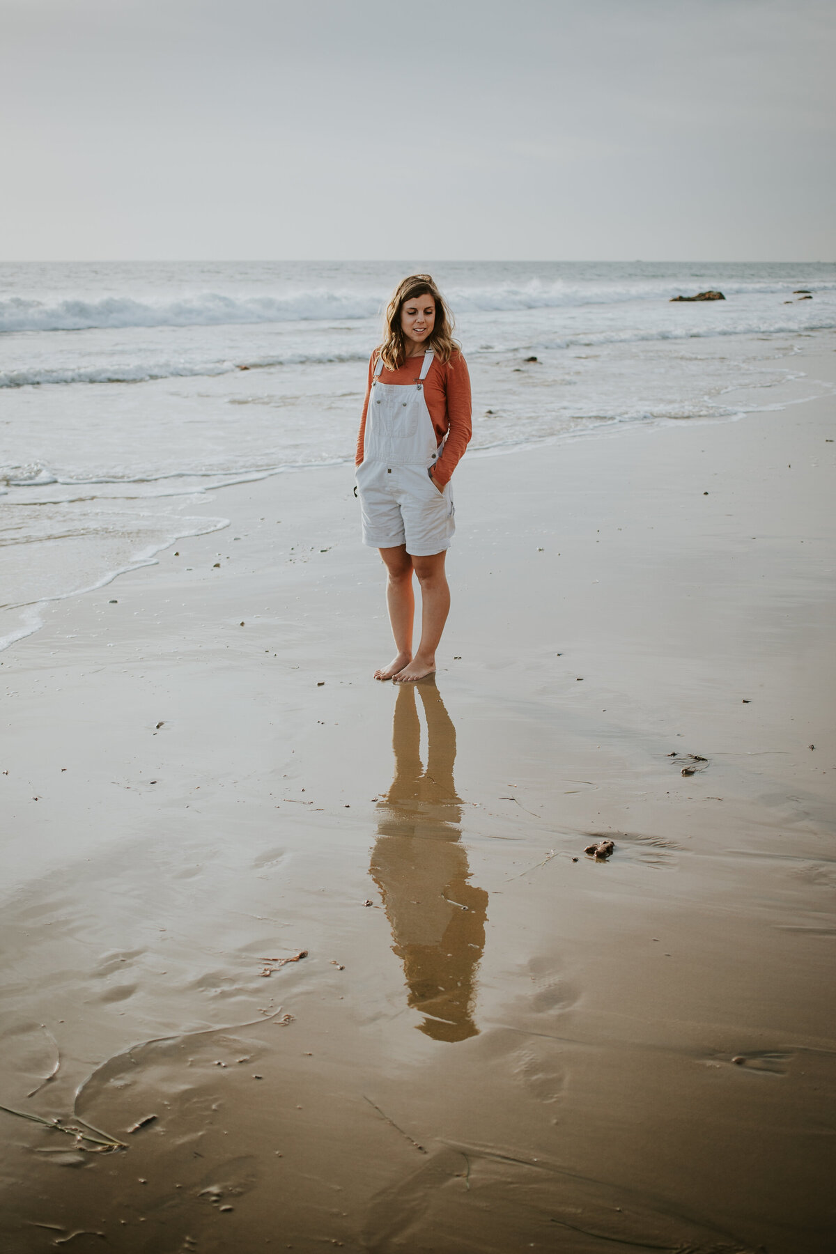 Young woman in overalls stands on beach while looking at reflection in wet sand.
