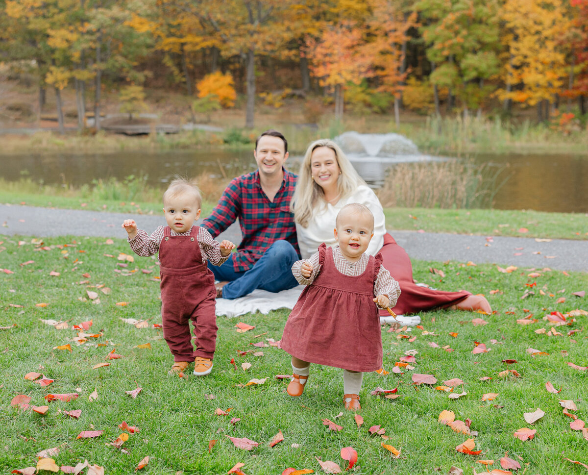 Central Massachusetts family photographer capturing twin toddlers walking toward the camera while their parents sit smiling by a pond surrounded by fall foliage