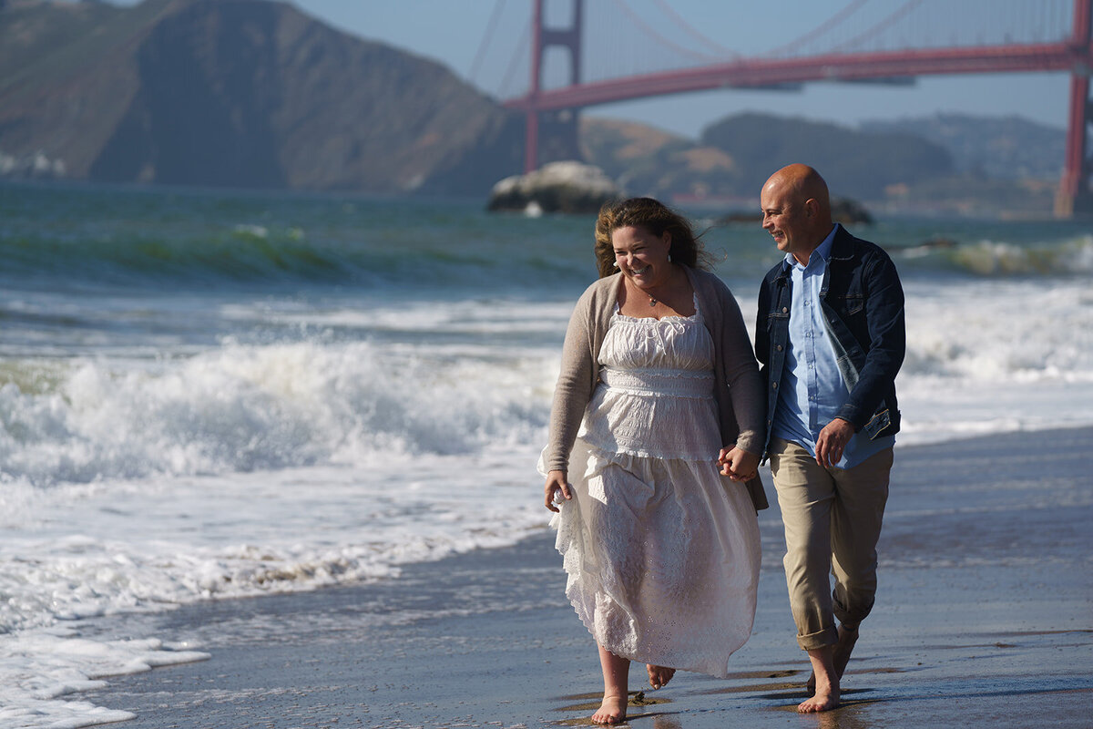 Engaged couple holding hands walking down the beach with the golden gate bridge in the background