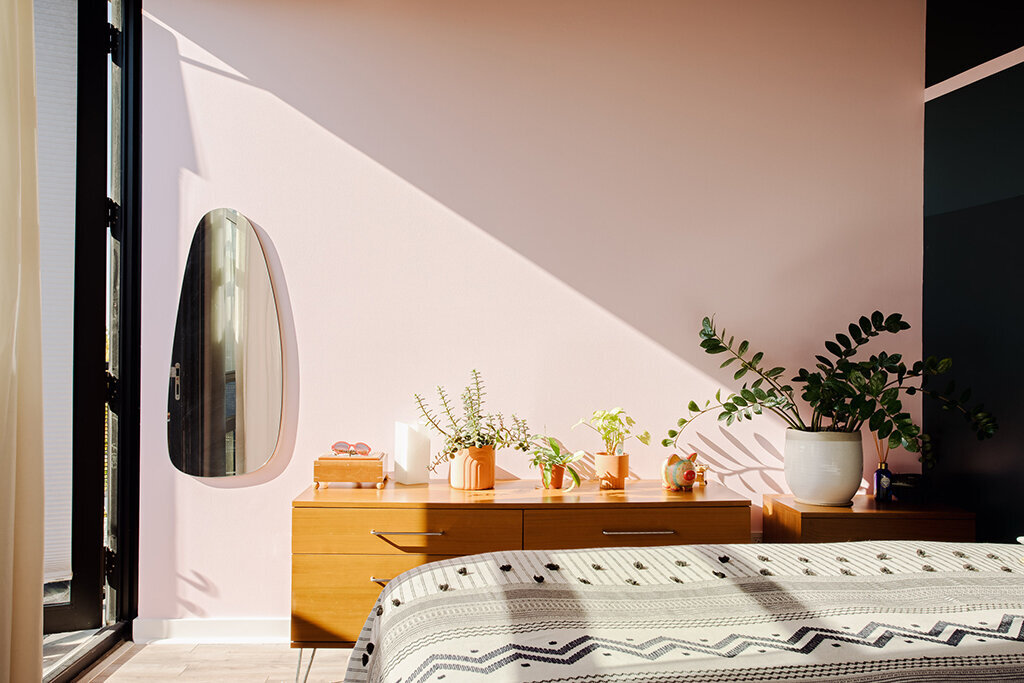 A sunlit bedroom with soft pink walls. A mid-century modern wooden dresser holds several small potted plants, a white candle, and a small wooden box. A large oval mirror hangs on the wall to the left of the dresser, reflecting light from a nearby window. A patterned white and gray duvet covers the bed in the foreground. A large potted ZZ plant casts shadows on the right wall.