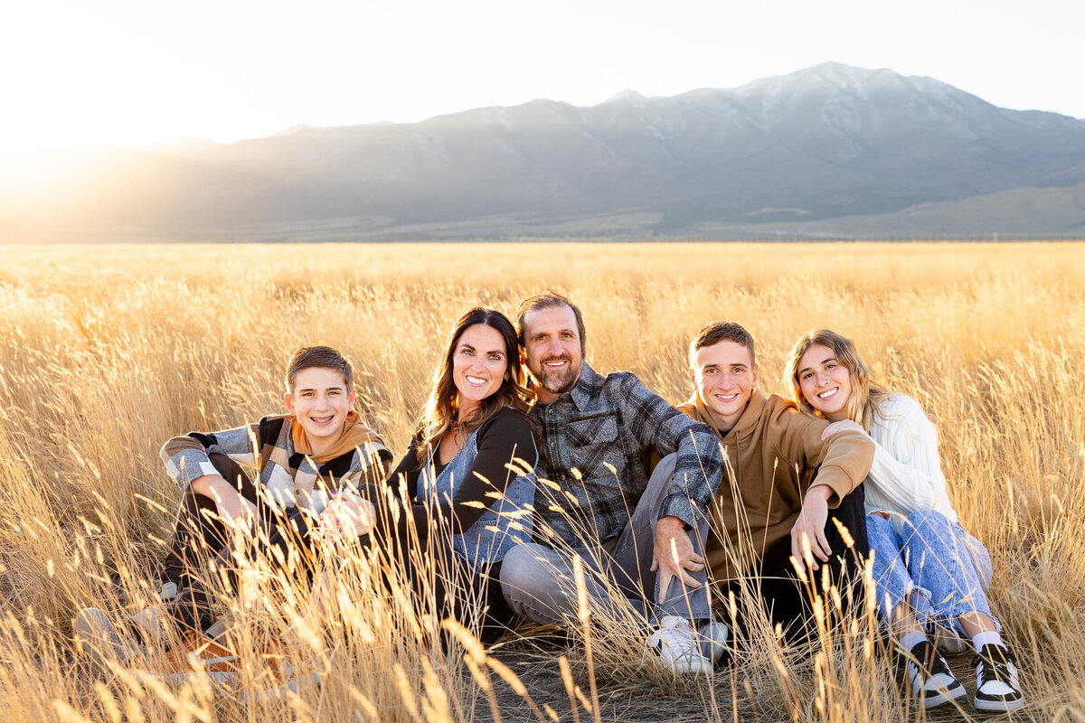  A family of five sits together in tall golden grass in Eagle Mountain, Utah, with the mountains in the background, all smiling warmly at the camera.