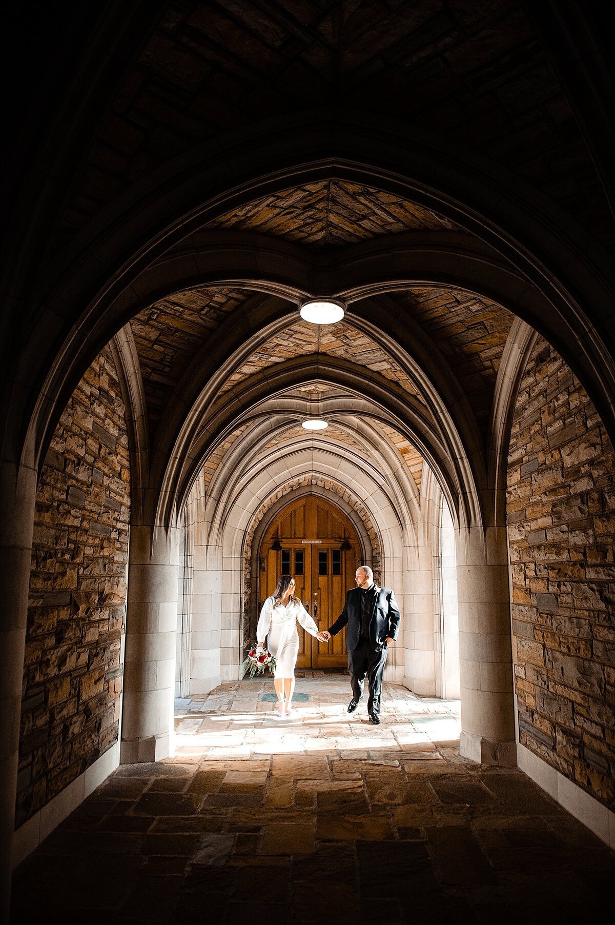 Standing in the arched entryway at Scarritt Bennett, the bride and groom hold hands as they walk. The bride is wearing a tea-length white dress with three quarter length sleeves, holding a pink, red and ivory bouquet. The groom is wearing a dark gray suit with a black shirt.