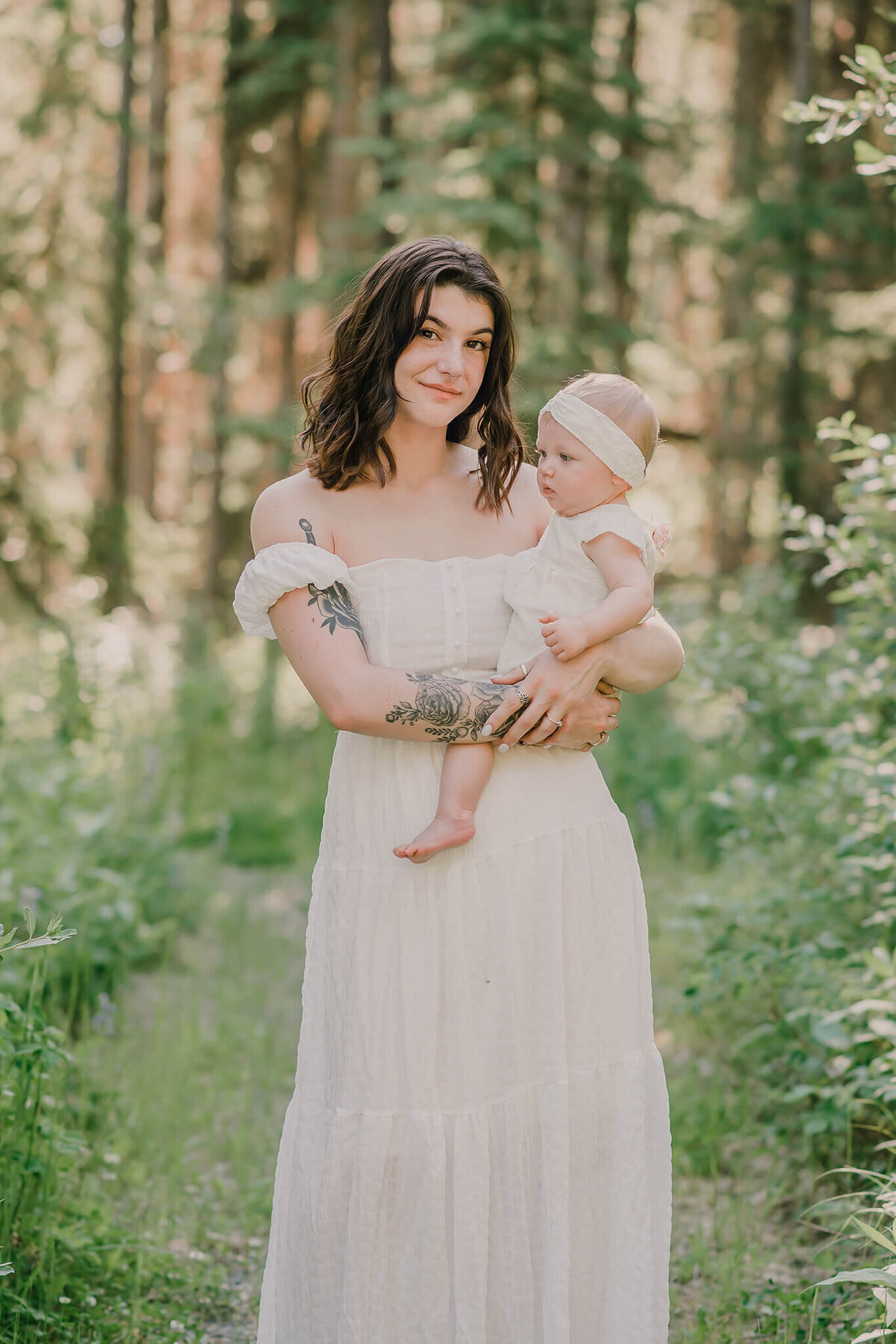 Mother holds her daughter on her hip in a lush green forest. Mother smiles at the camera while baby girl looks off into the distance.  Portrait by Kamloops Photographer Brittany Danielle