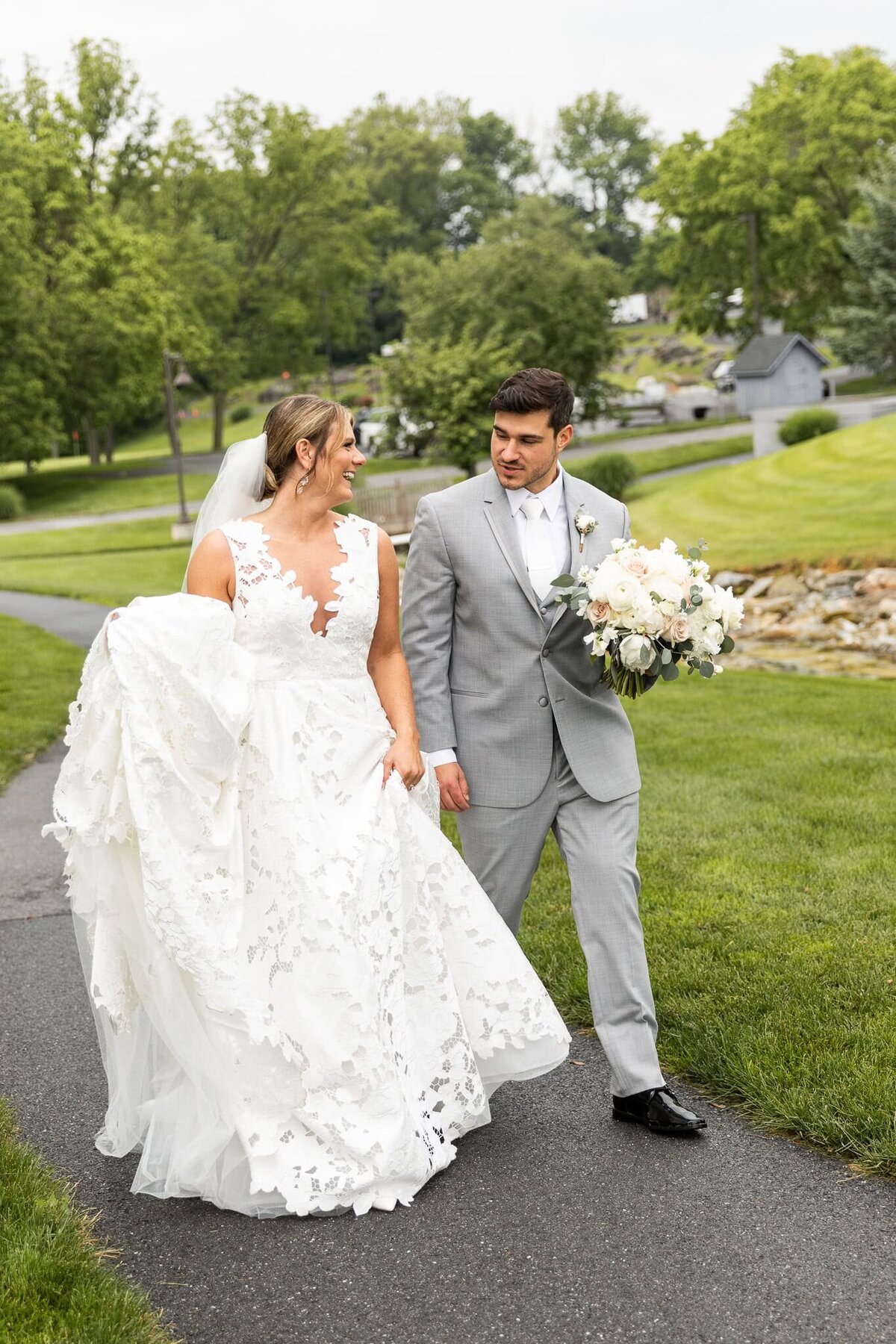  Newlyweds stroll through the Pennsylvania countryside as the bride lifts the hem of her intricate lace wedding gown as she walks hand in hand with her groom, who carries a lush bouquet of white roses and greenery. His light gray suit complements the soft, rolling hills behind them