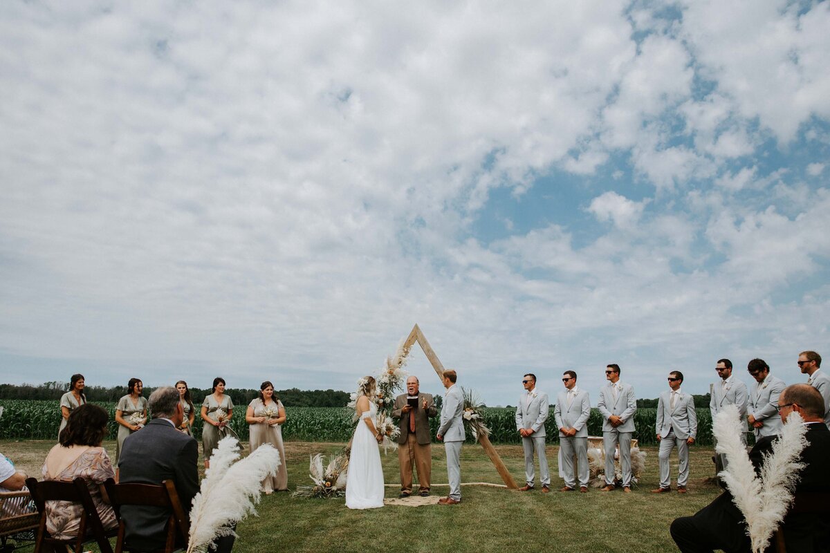 Bride and groom exchanging vows at an outdoor wedding ceremony in Exeter, Ontario. They are standing in front of a triangular wood arbour backdrop with flowers.