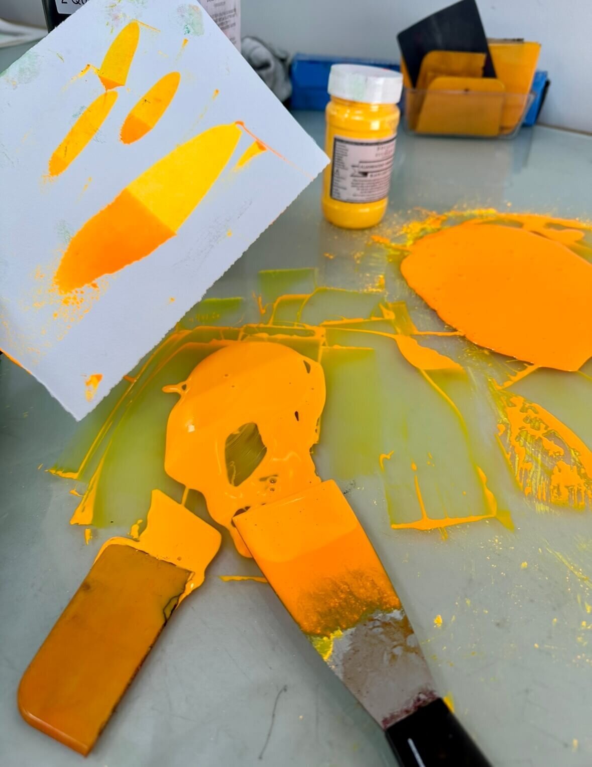 A vivid yellow-orange ink in progress, with test swatches, palette knives, and pigment jars scattered across a glass mixing surface.