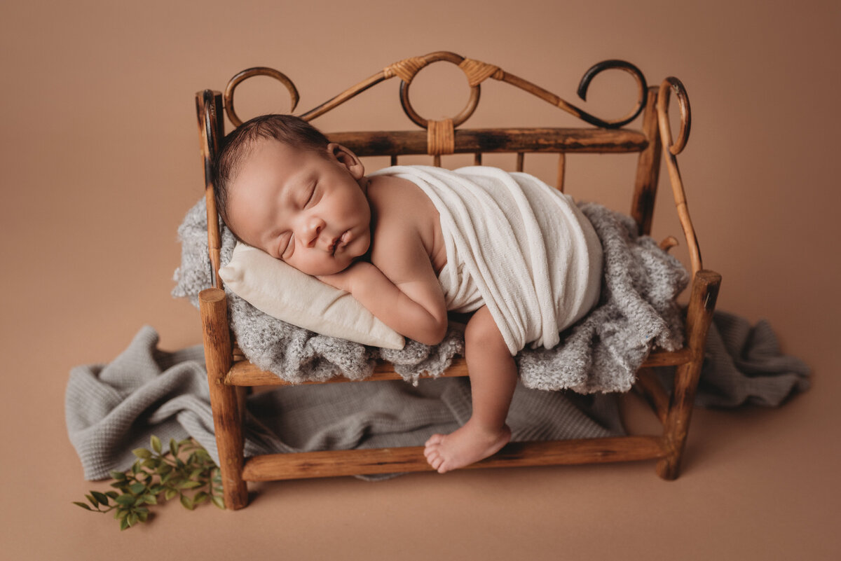Newborn boy asleep on stomach laying on mini bench and tiny pillow with white wrap around his bottom on tan backdrop with greenery under bench