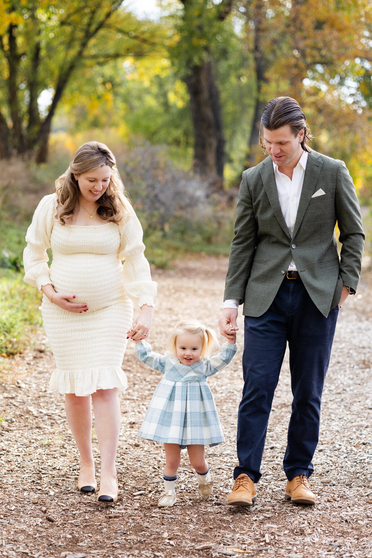 Toddler girl holds her mom and dad's hands as they smile at her and they all walk down a wooded path.