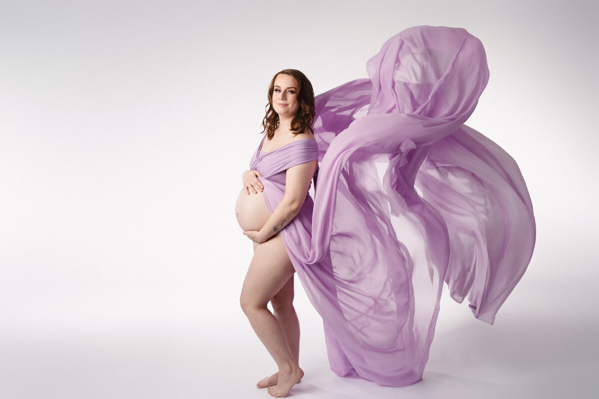 Pregnant mother to be wearing a purple maternity gown holding her tummy as the fabric of her gown blows behing her