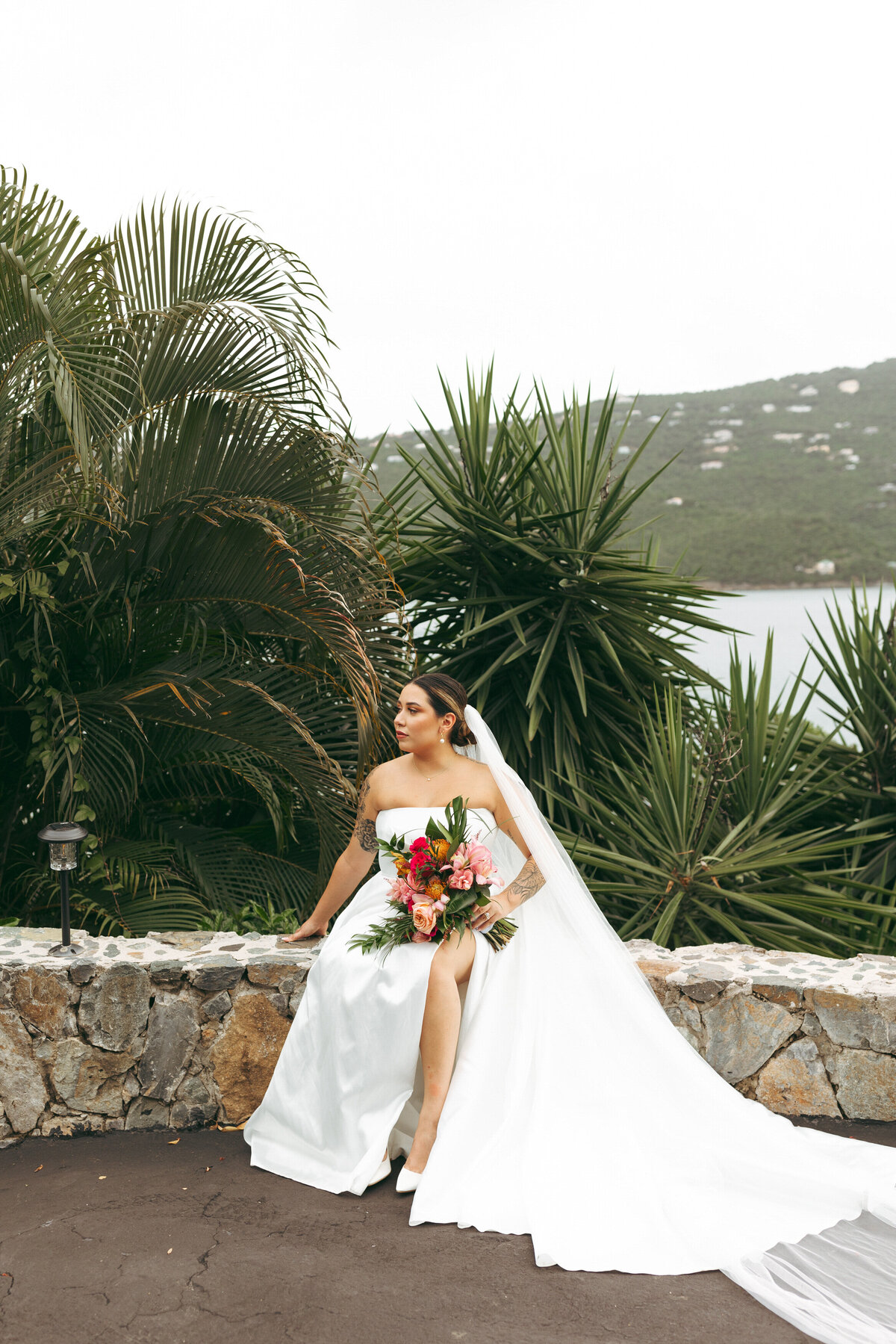 Elegant bride sitting on a stone wall with lush tropical plants in the background