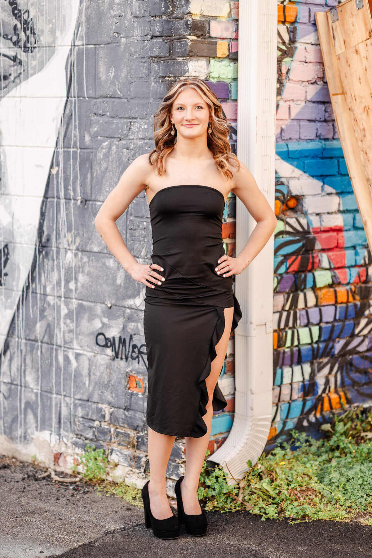 A high school senior in a black dress puts her hands on her hips and smiles. Behind her, you can see two walls with murals; one colorful and one monotone. This is the full body pull back of another image.