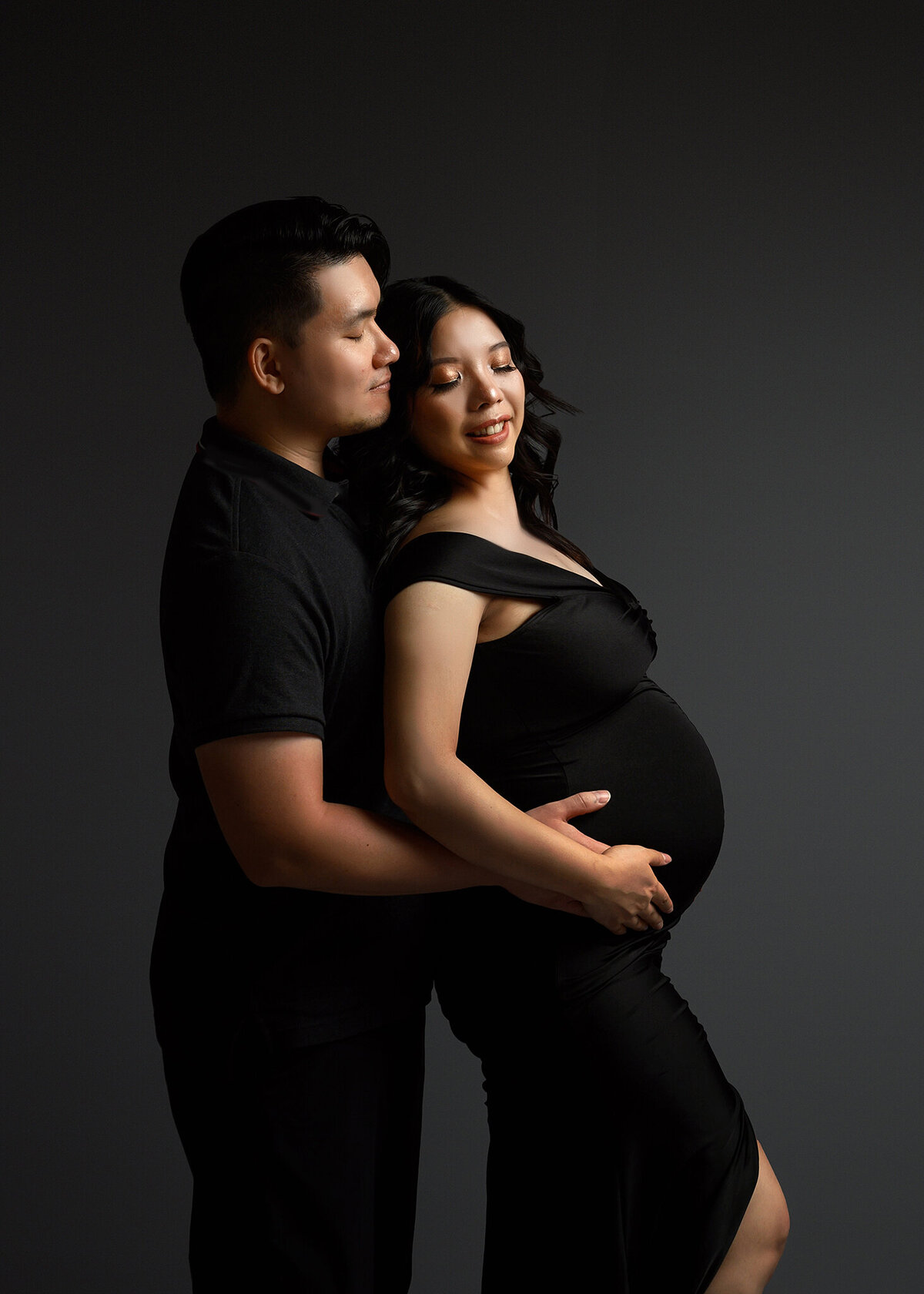 Simple photo of husband embracing pregnant wife with both of their hands on the belly, wearing black formal attire, simple dark gray background in a Phoenix Maternity studio session