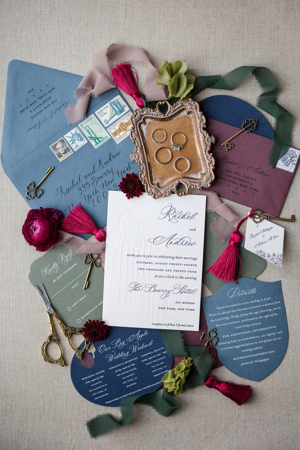 Wedding invitation suite with cards in various colors, gold rings on a tray, tassels, stamps, and decorative scissors, arranged on a neutral fabric background. This photo was taken by a NYC wedding photographer for rooftop ceremonies