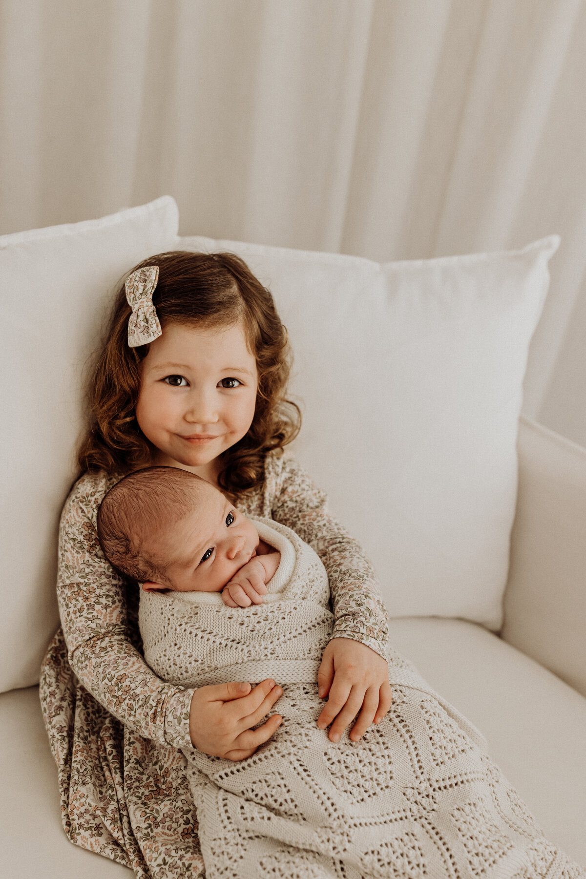 A little girl holds her newborn baby brother sitting on a white sofa that could be in anyones home, photographed in christchurch by lucy jane