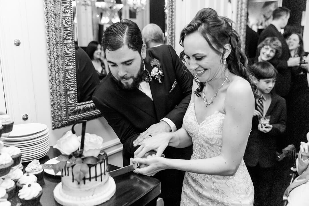 A bride and groom laugh while cutting their cake with a large knife