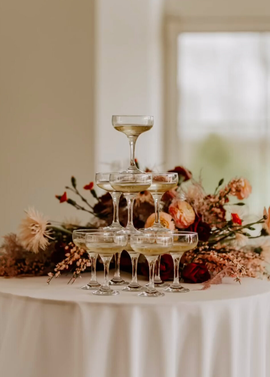 Structure à trois étages de coupes de champagne disposés sur une table avec une nappe blanche et immense bouquet de fleurs derrière. photo prise par Laura, photographe de mariage en vendée.