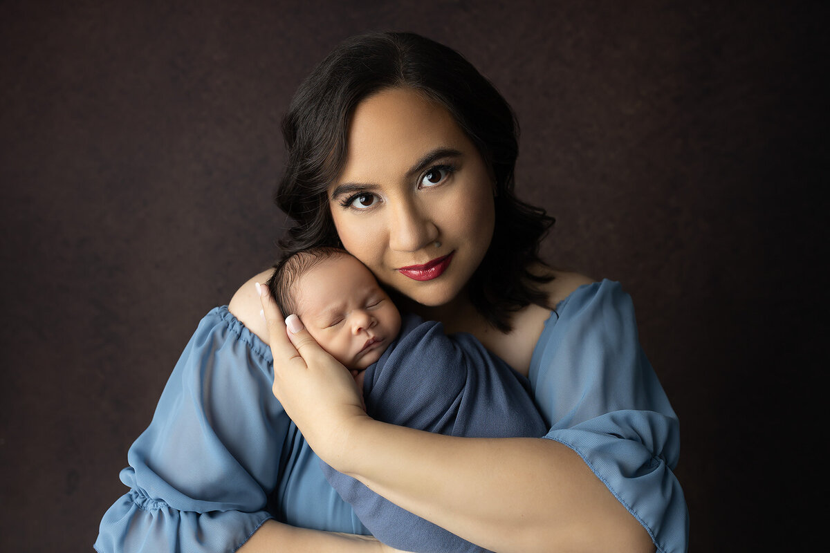 New mother lovingly holding her swaddled newborn baby in a soft blue dress during their Dallas newborn photography session.