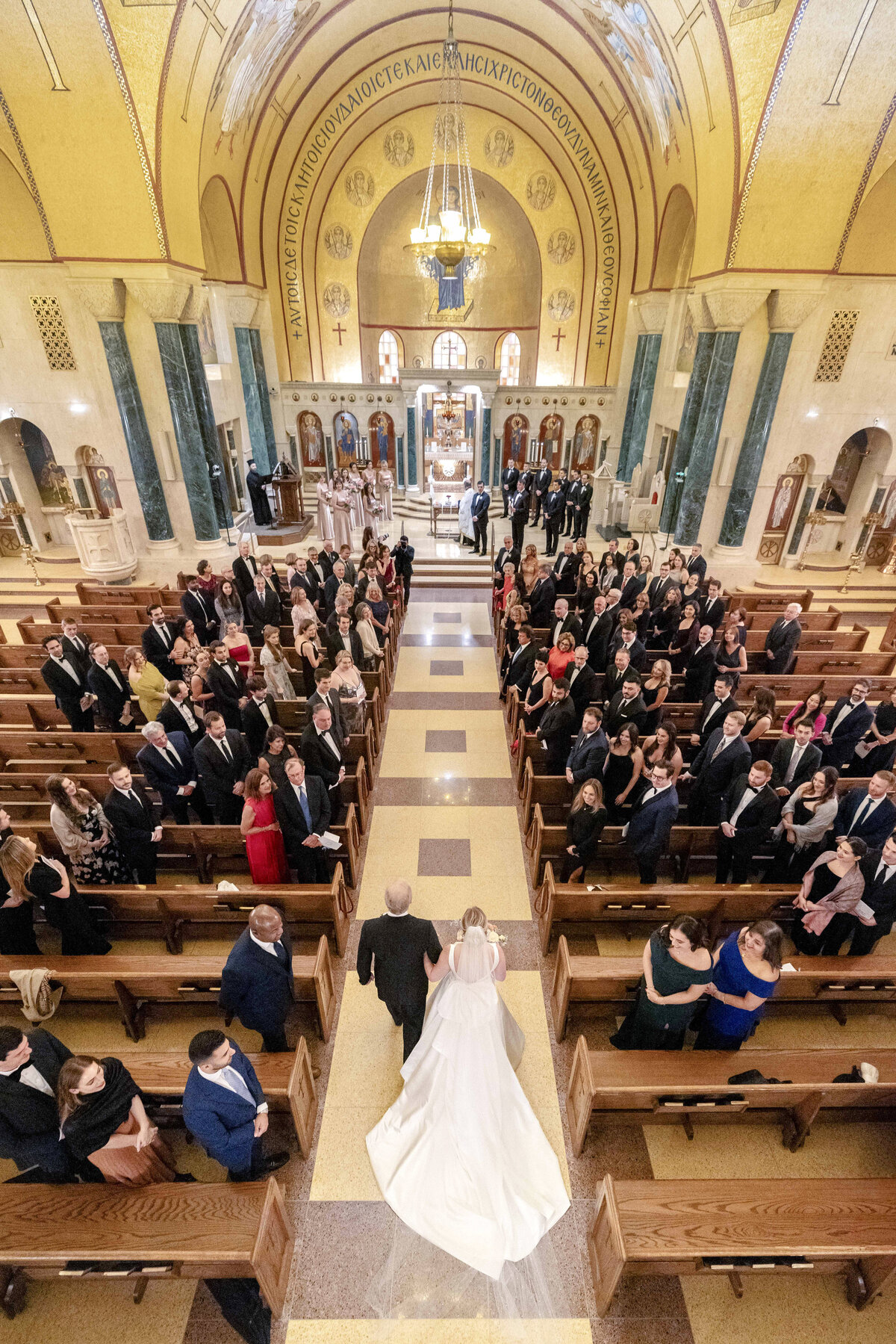 A bride walks down the aisle of a grand church, accompanied by an older gentleman. The guests are seated on either side, with some standing and looking on. The church features ornate gold and green decorations and a high arched ceiling.