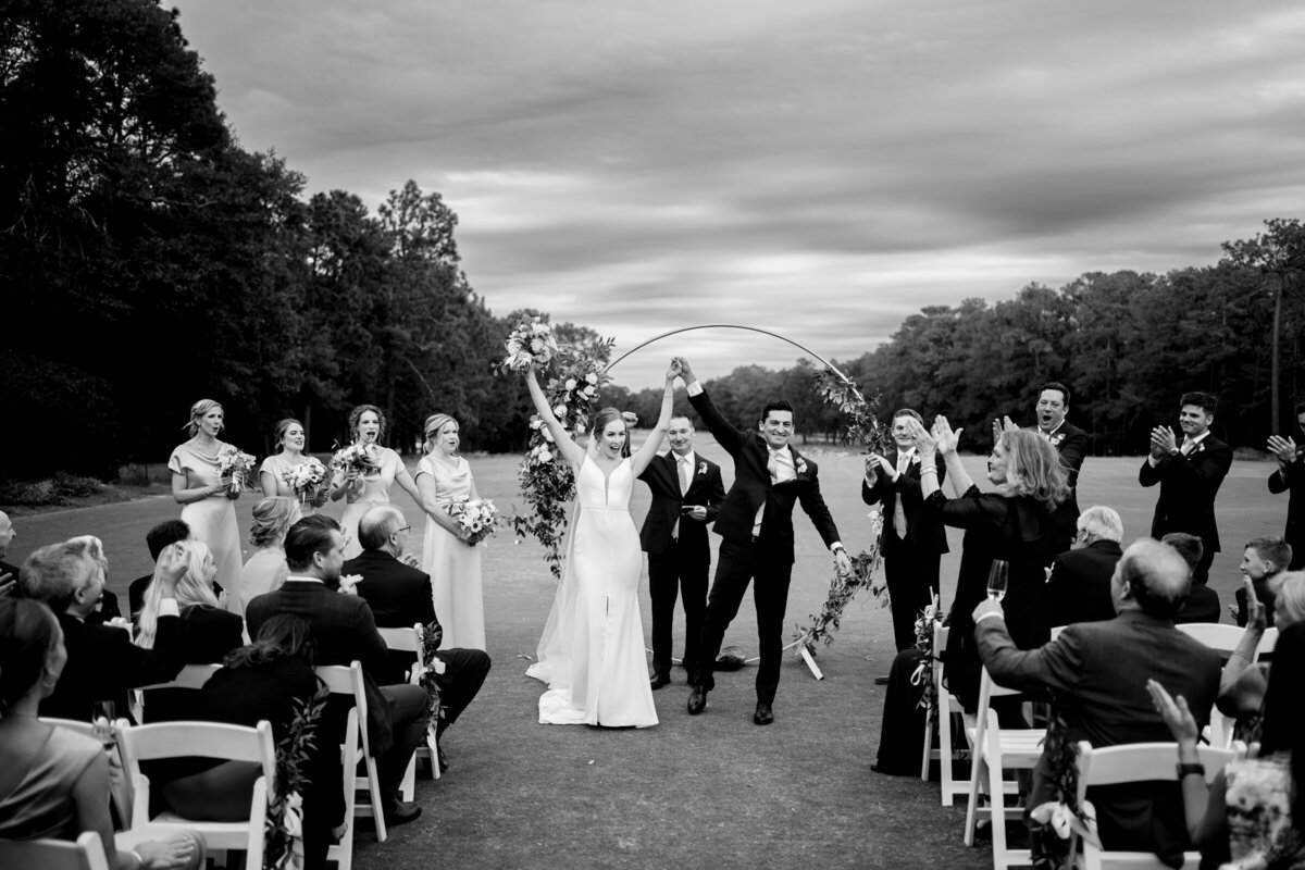 A joyous couple raises their hands in victory as they walk down the aisle, surrounded by applauding guests in a black and white photo.